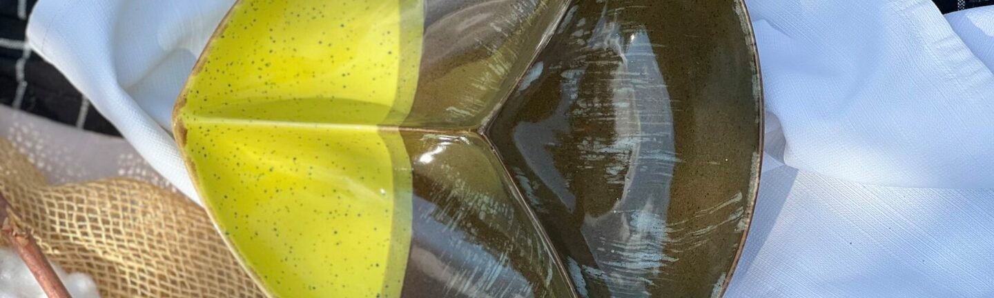 Ceramic plate in a triangular shape with two colors: yellow and dark green, placed on a white towel, with a background of cotton branches and fabric mesh.