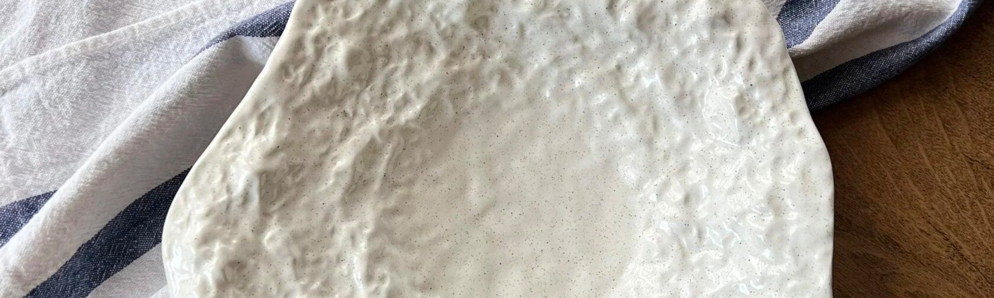 A ceramic plate with an unusual organic shape and a textured white surface, placed on a wooden table with cloth napkins nearby. The background is decorated with a branch with cotton flowers.